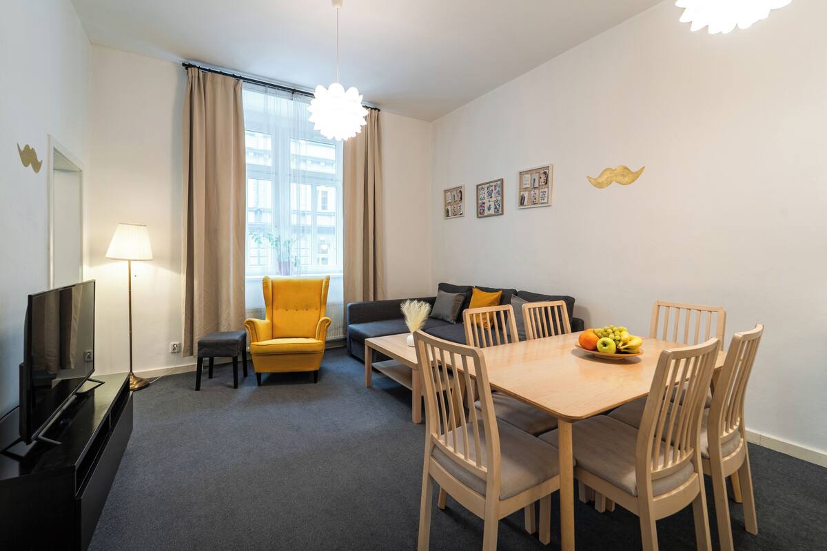 Uma sala de estar moderna com um sofá, uma poltrona amarela, uma mesa de jantar com cadeiras e uma televisão de tela plana.