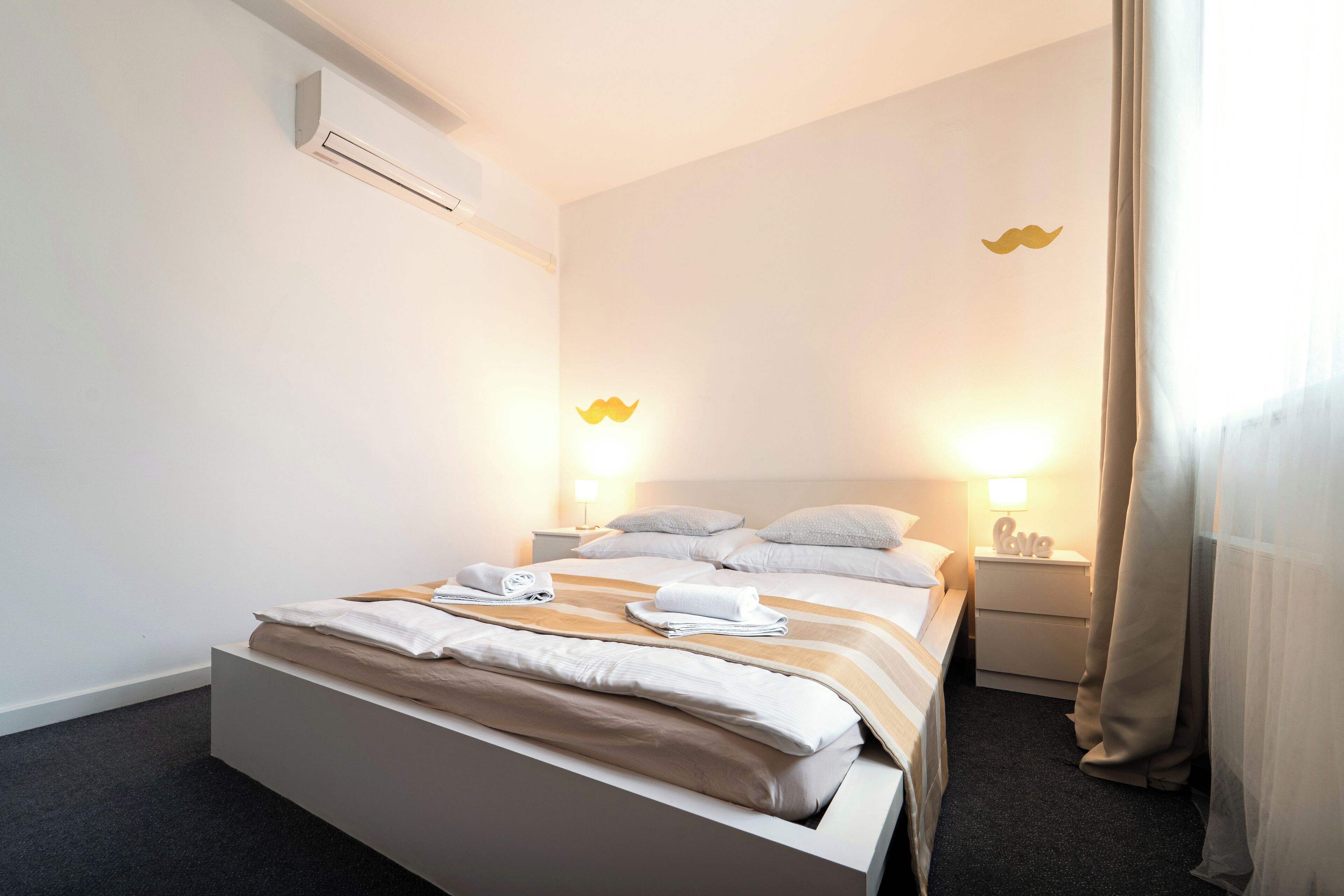 A neatly made bed with a white and beige striped blanket, a small bedside table with a lamp, and a wall-mounted air conditioner.