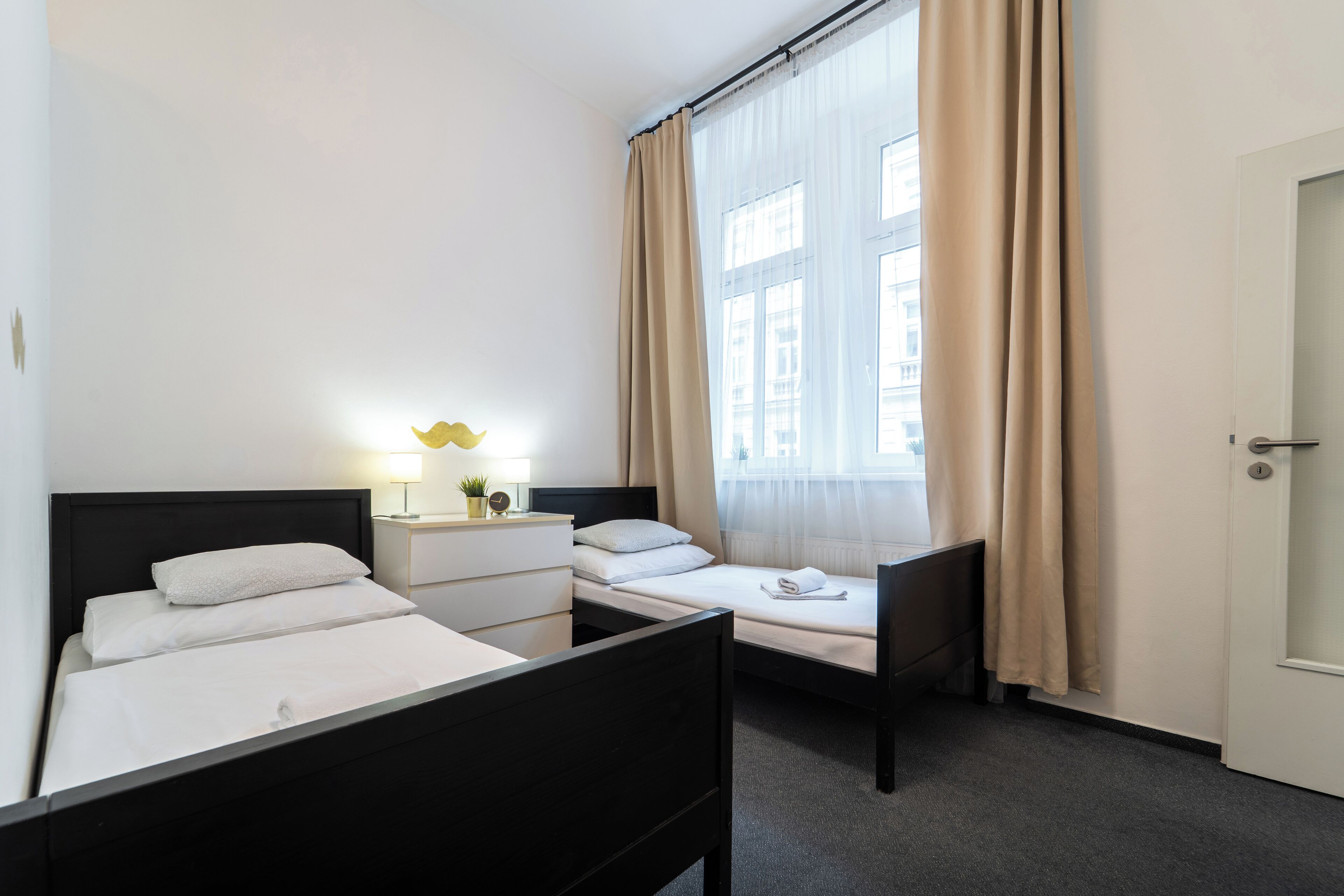A hotel room with two beds, a bedside table with a lamp, a chair, and a door.