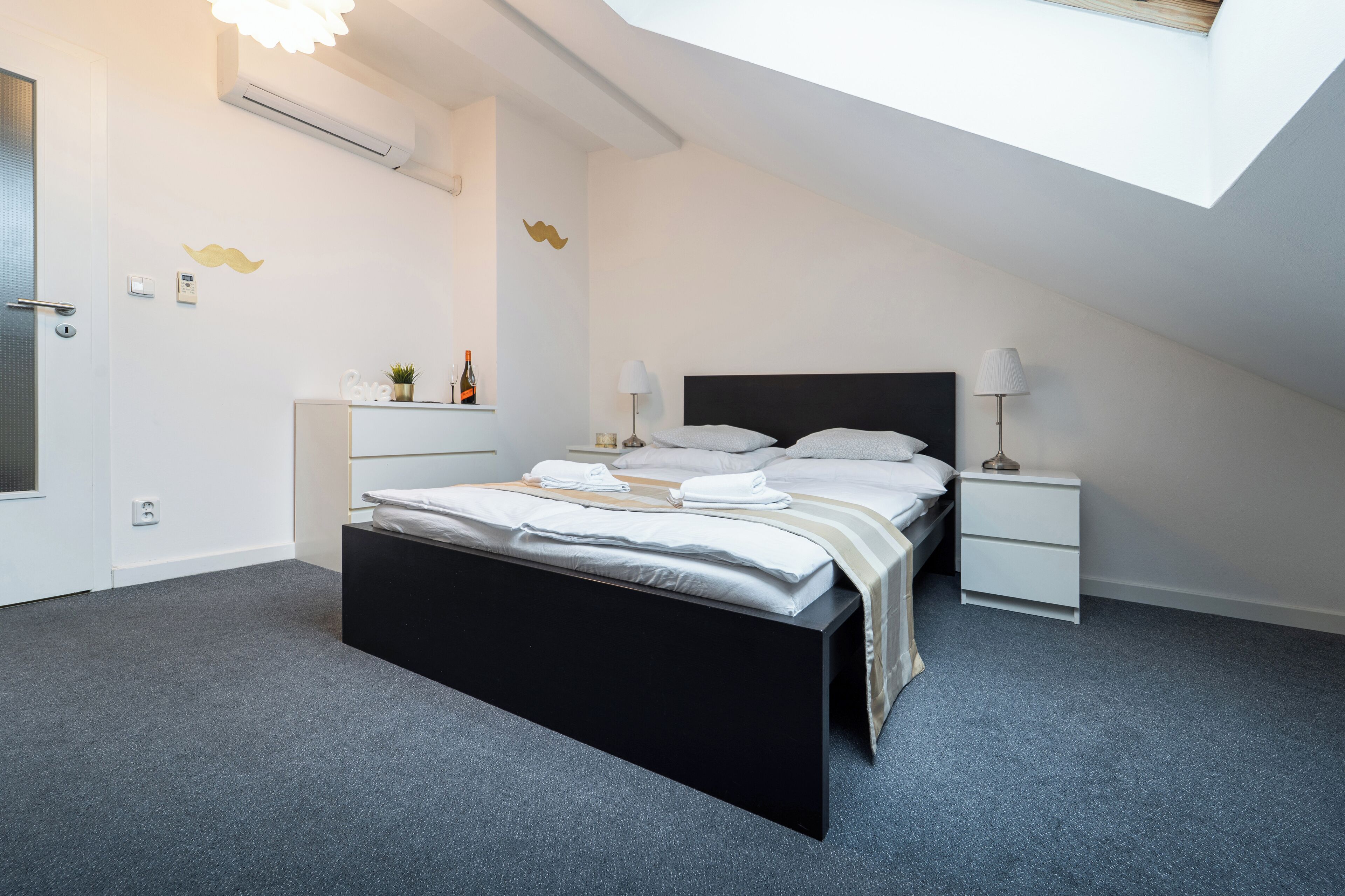 Un dormitorio moderno con estructura de cama negra, ropa de cama blanca, una mesita de noche pequeña con una lámpara y un aire acondicionado de pared.