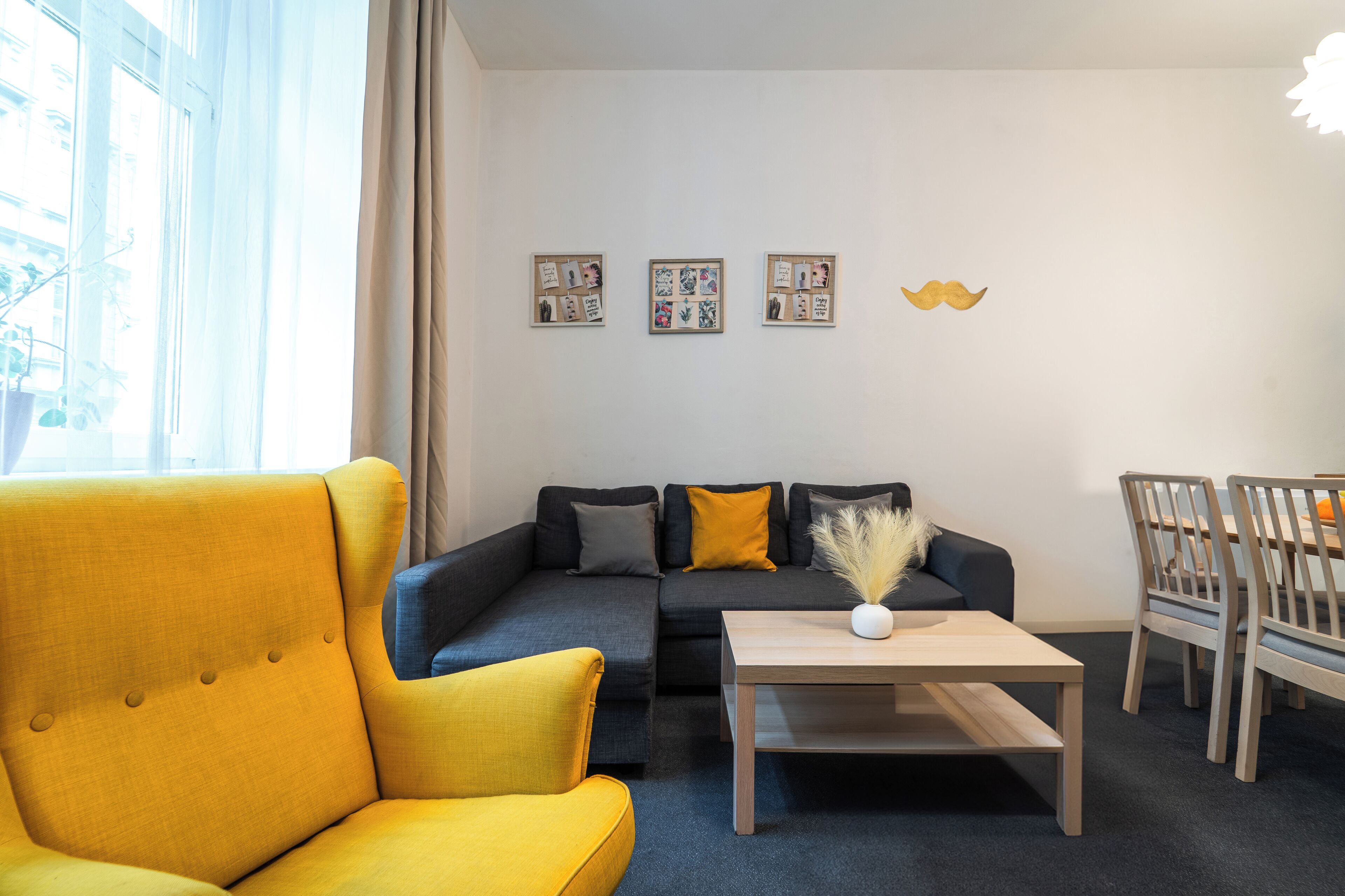 A modern living room with a yellow armchair, a dark sofa, a wooden coffee table, and a dining area with wooden chairs and a table.