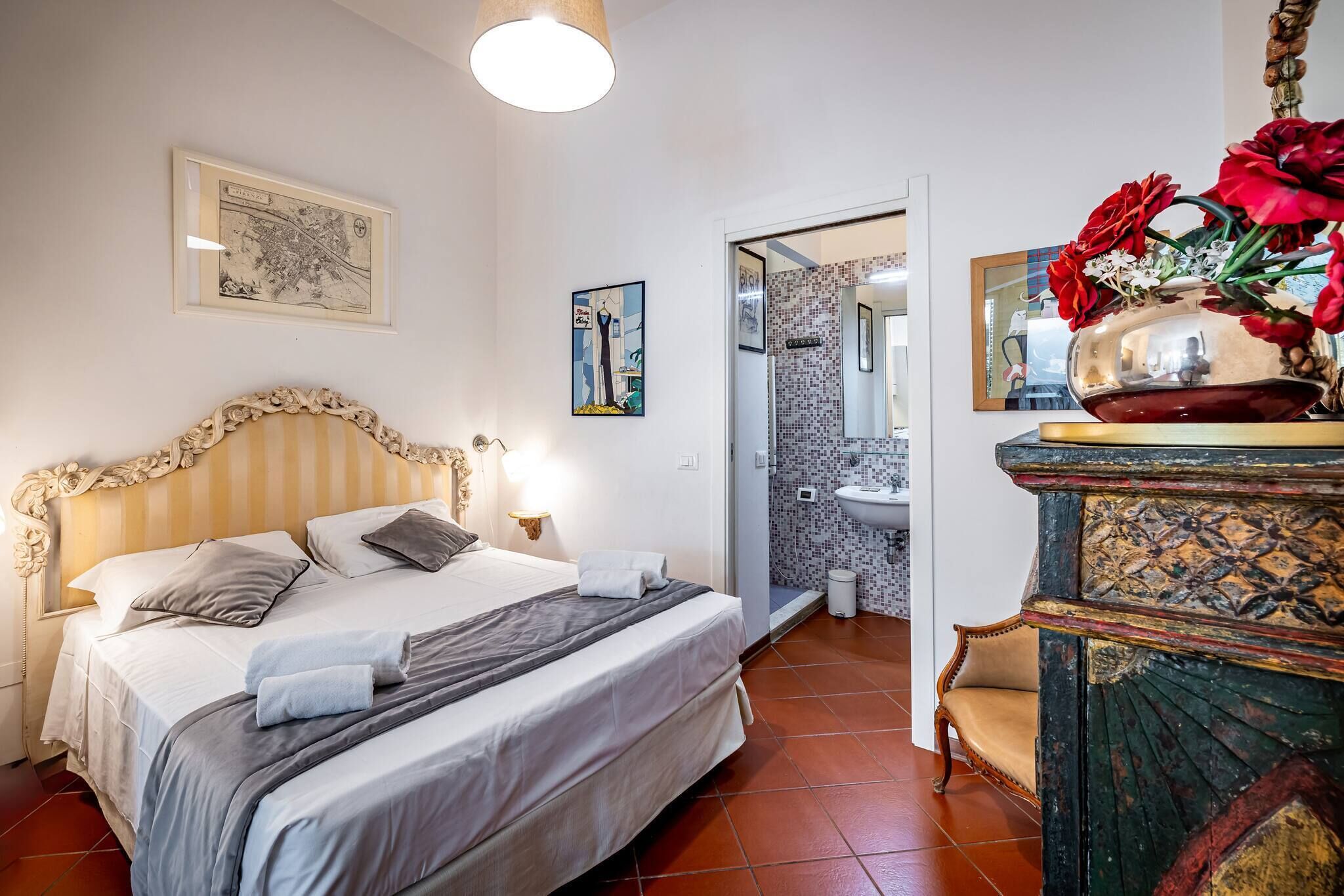 A hotel room with a bed, a nightstand, a mirror, a vase with flowers, and a bathroom visible through an open door.