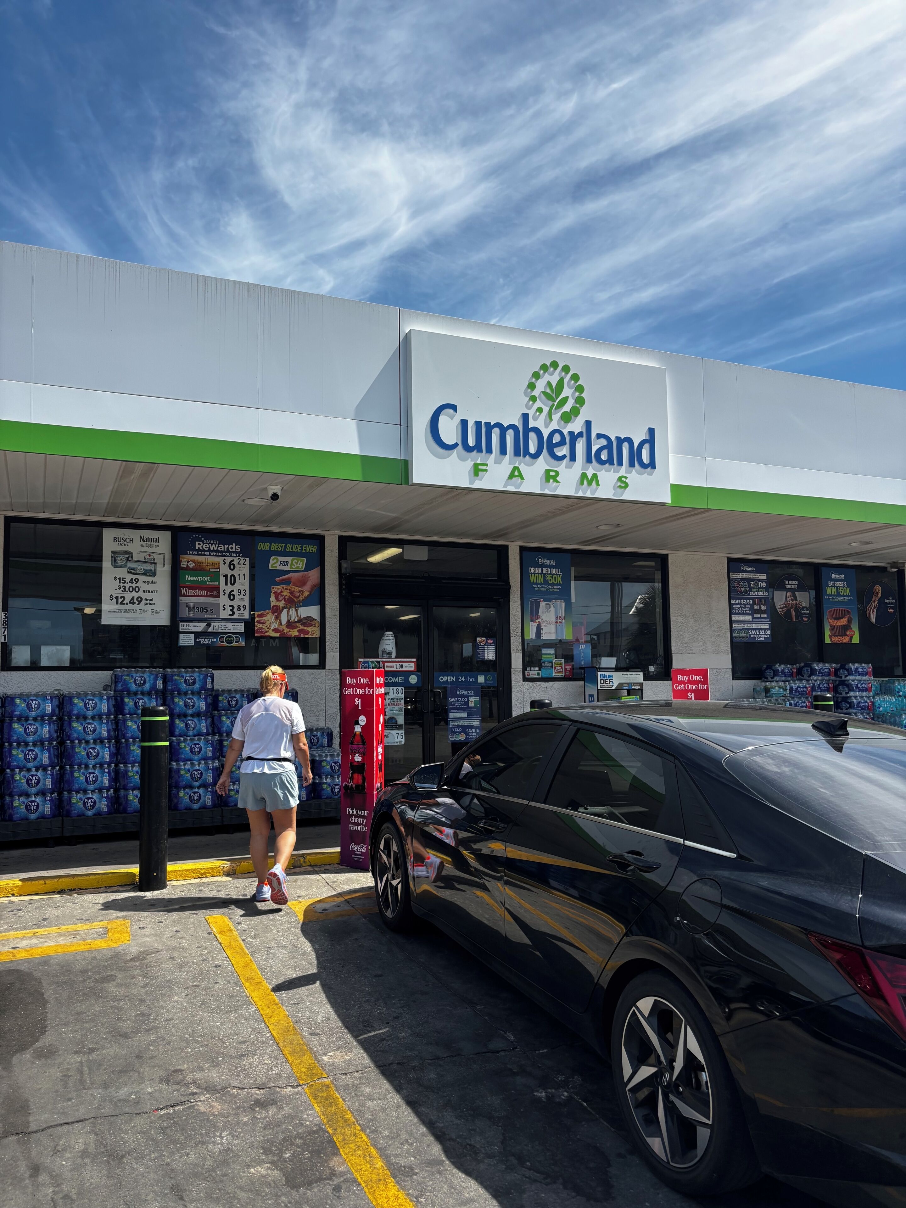 Una tienda Cumberland Farms con fachada verde y blanca, un coche negro estacionado adelante y botellas de agua apiladas afuera.