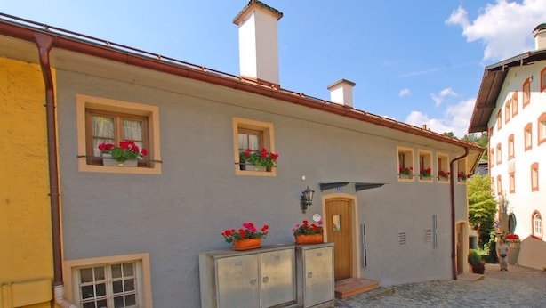 A building with a light gray facade, multiple windows with flower boxes, a wooden door, and a cobblestone courtyard. - Apartment Alte Fronfeste II 40sqm (Berchtesgaden)