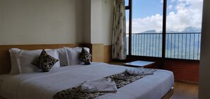 A hotel room with a large bed, a small bedside table, and a view of a mountain through a balcony railing.