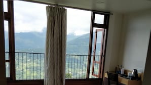 A hotel room with a large bed, a small table, a bench, a desk with a chair, and a view of mountains through large windows. - Hotel Ninamma (Gangtok)