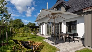 Een terras met een tafel en stoelen, een grote parasol en een gazon met twee lounge fauteuils.