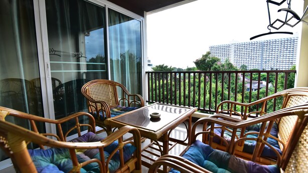 A balcony with wicker furniture and a glass table, overlooking a residential area and a tall building. - Joy House (Pattaya)