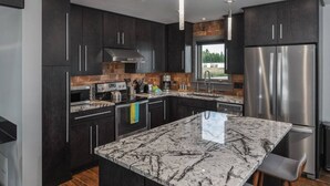 Una cocina moderna con armarios oscuros, una isla grande con encimera de mármol, electrodomésticos de acero inoxidable e iluminación colgante.