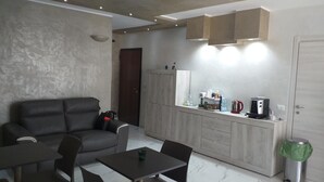 Una sala de estar moderna con un sofá gris, una mesa de comedor y una cocina equipada con microondas, hervidor y cafetera.