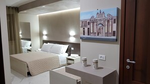 Habitación de hotel con cama, mesitas de noche, escritorio, espejo y un cuadro de un edificio.