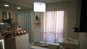 Una cocina moderna con barra desayunadora, zona de comedor con mesa y sillas, y un gran ventanal con cortinas ligeras.