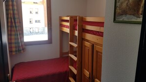 A small, compact room with a bunk bed, a single bed, a window with curtains, and a framed picture on the wall.