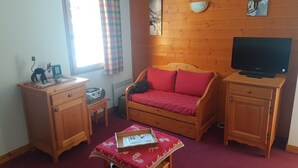 A small, cozy room with a wooden bed, a television, a desk with a radio, and a window with curtains.