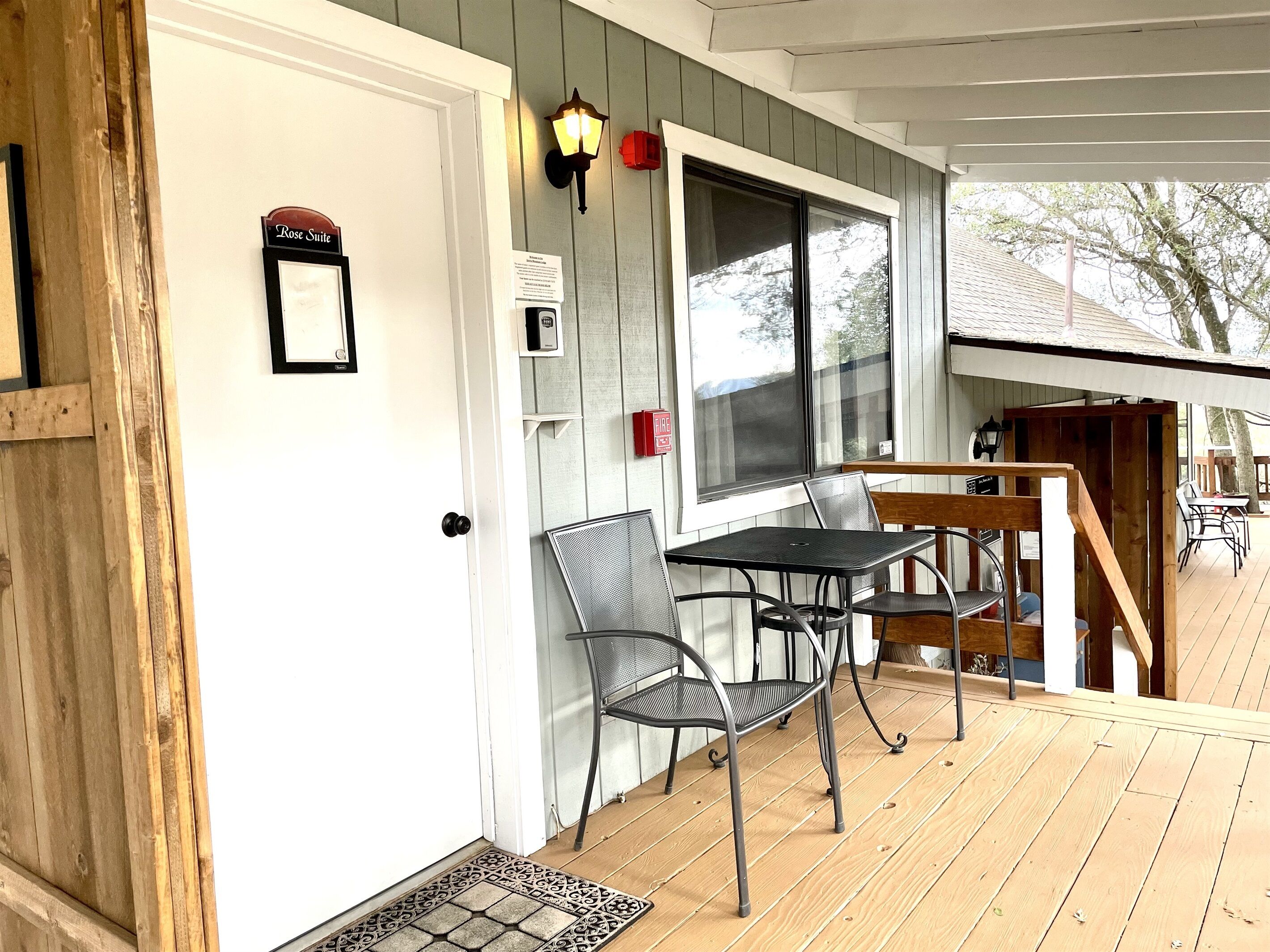 A covered outdoor seating area with a table and chairs, a door with a sign, and a window with a view of trees.