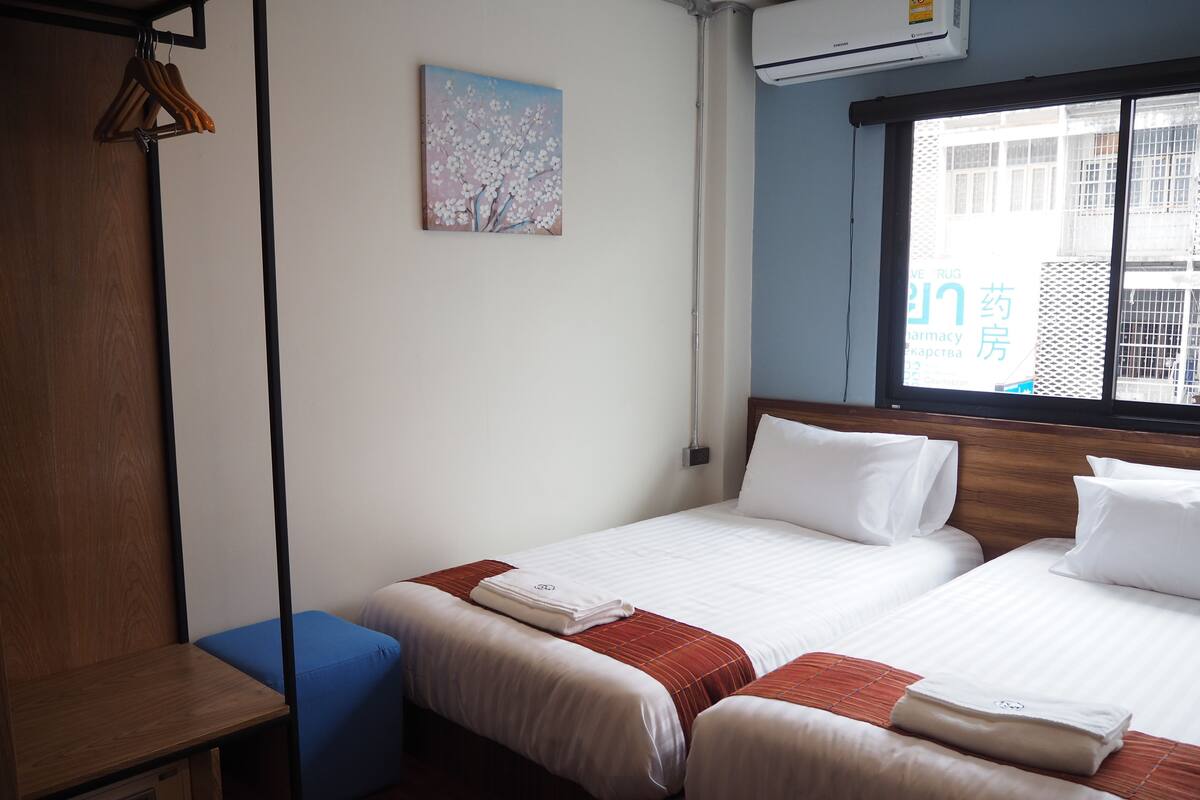 A hotel room with two beds, a wooden wardrobe, a blue bench, a painting on the wall, and a window with a view of buildings.