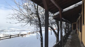Uma área externa coberta com chão coberto de neve, vigas de madeira e vista para uma paisagem nevada.
