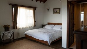 Quarto com cama de madeira, mesa de cabeceira com um vaso de flores, radiador e janela com cortinas.