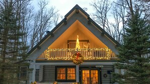 Eine Blockhütte mit Balkon, geschmückt mit Lichtern und Girlanden, umgeben von Schnee und Bäumen.