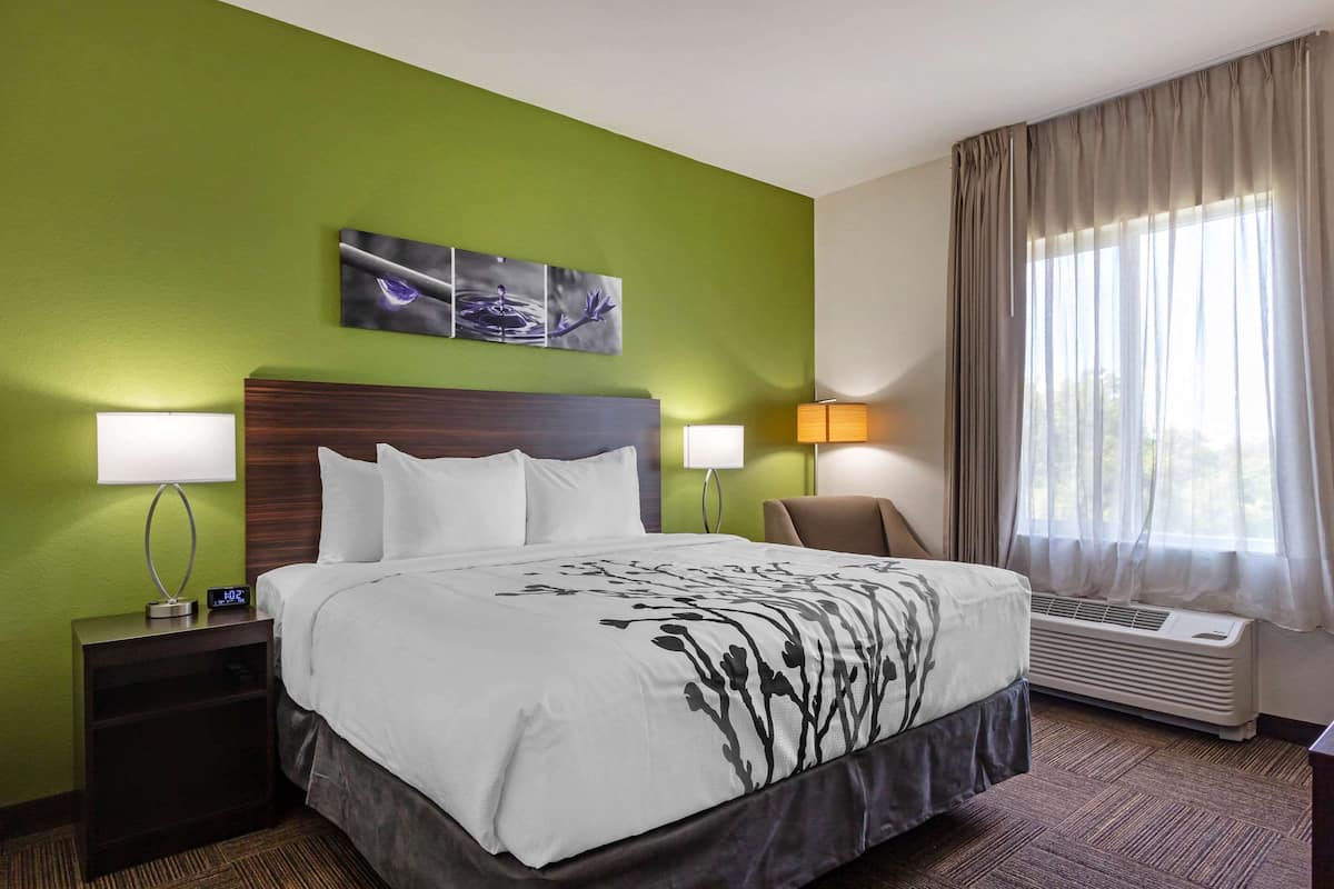A hotel room with a large bed, two bedside lamps, a chair, a wall-mounted air conditioner, and a window with curtains.