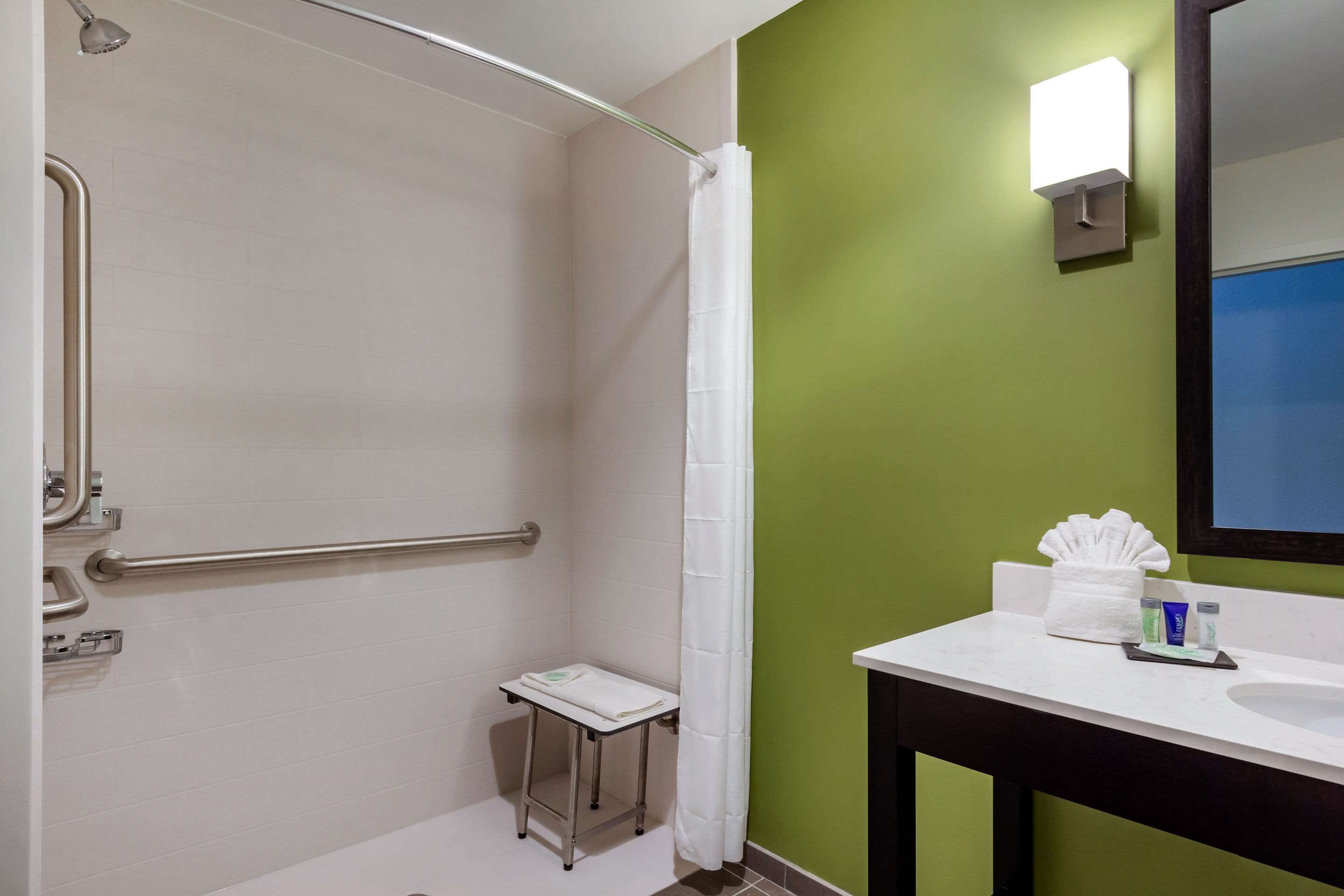 A bathroom with a shower, grab bar, small step stool, and a sink with toiletries.