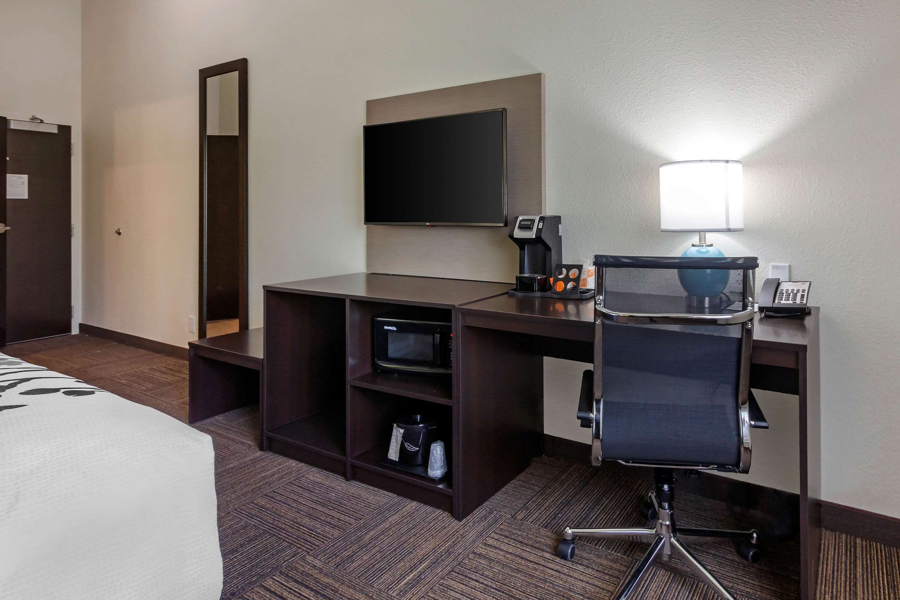 A hotel room with a bed, a desk with a computer, a chair, a flat-screen TV, a coffee maker, and a lamp.
