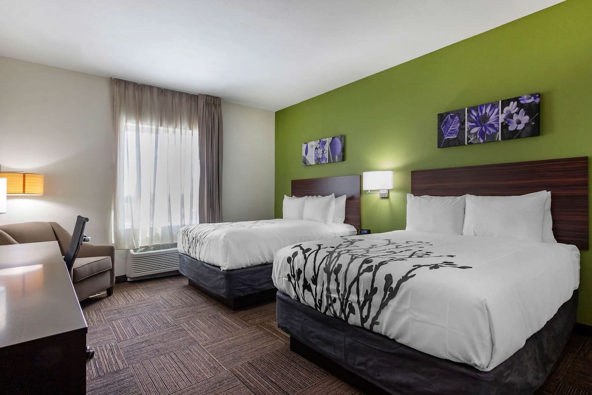 A hotel room with two beds, a desk, a chair, a lamp, and a window with curtains.