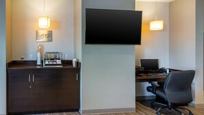 Ein Hotelzimmer mit dunklem Holztisch, einem an der Wand befestigten Flachbildfernseher, einem bequemen Bürostuhl und einer Minibar mit verschiedenen Artikeln.
