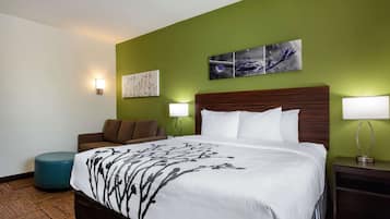 Una habitación de hotel con una cama grande, un sillón marrón, un otoman teja y dos lámparas de pared.