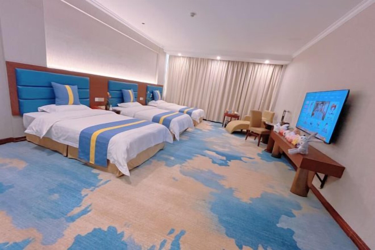 Habitación de hotel con tres camas, un televisor de pantalla plana, un banco de madera y un ventanal con cortinas.