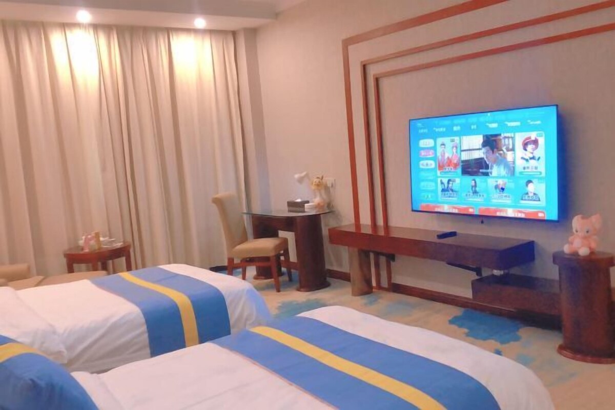 Habitación de hotel con dos camas, un televisor mostrando una videollamada, un escritorio con una silla y una mesita de noche con una lámpara.