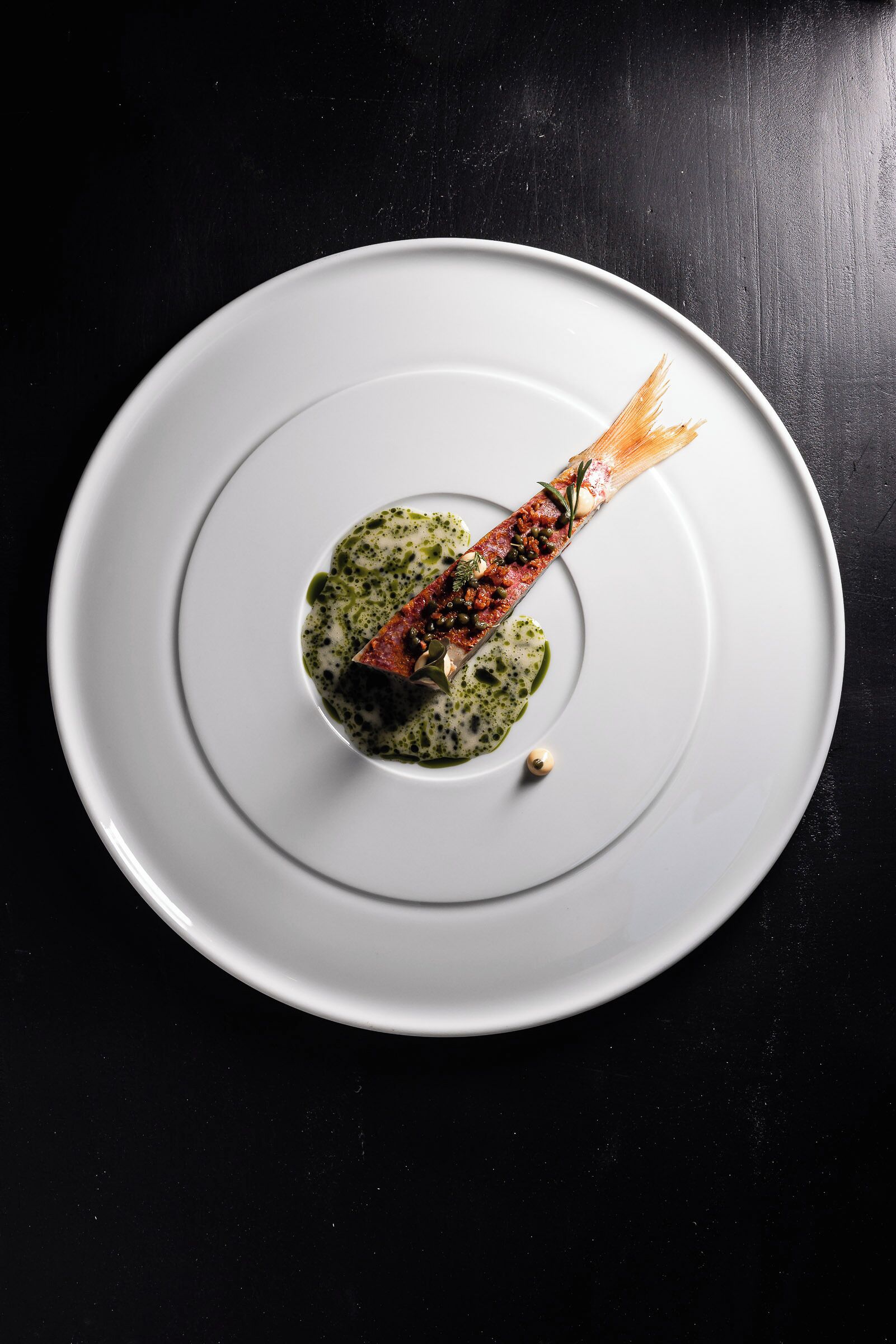 Un plato blanco con pescado y salsa verde sobre una superficie negra.