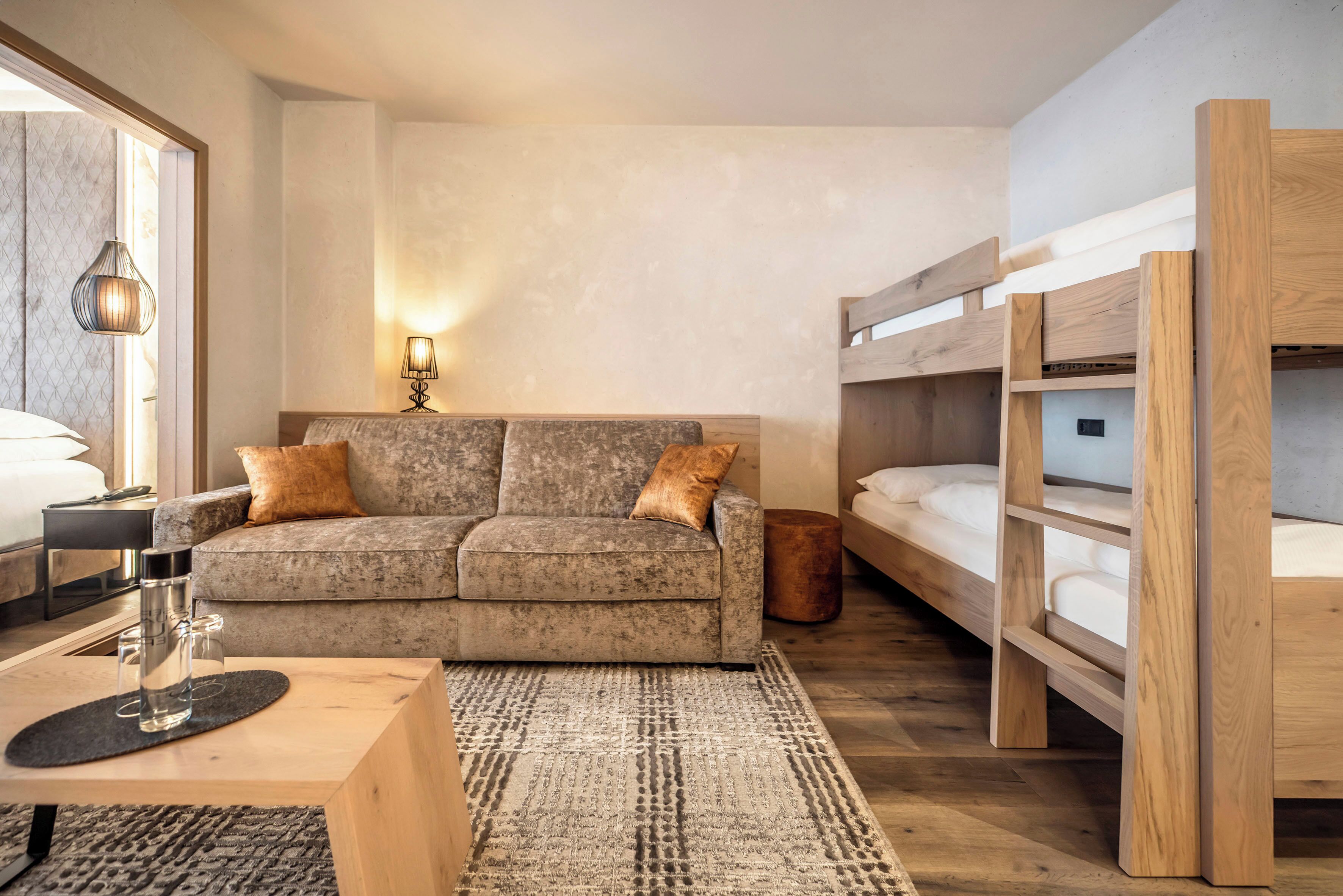 Une chambre moderne avec un canapé gris, un lit superposé en bois, une petite table surmontée d’un récipient en verre et un tapis à motifs.