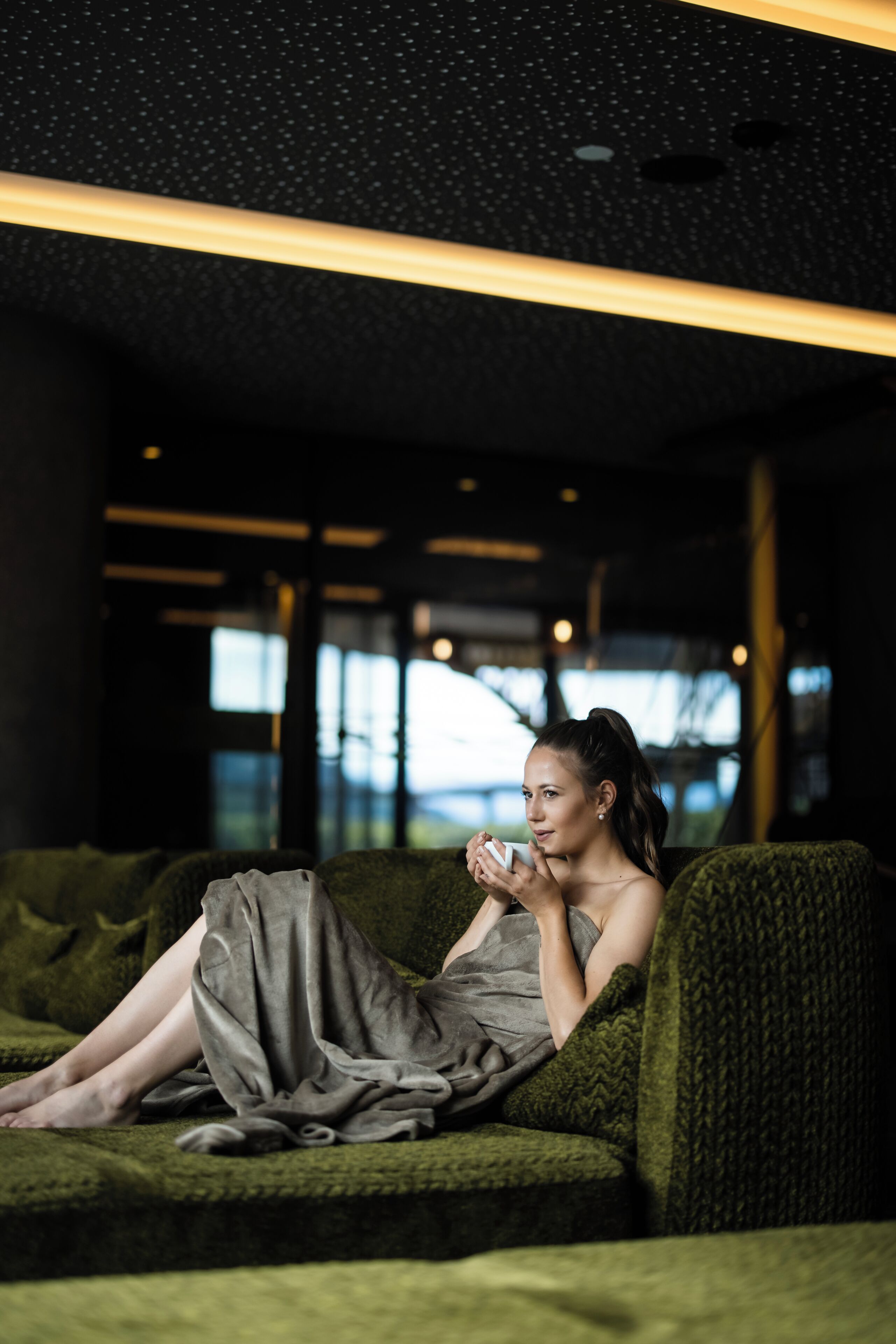 A person sitting on a green sofa in a modern interior with a dark ceiling and large windows.