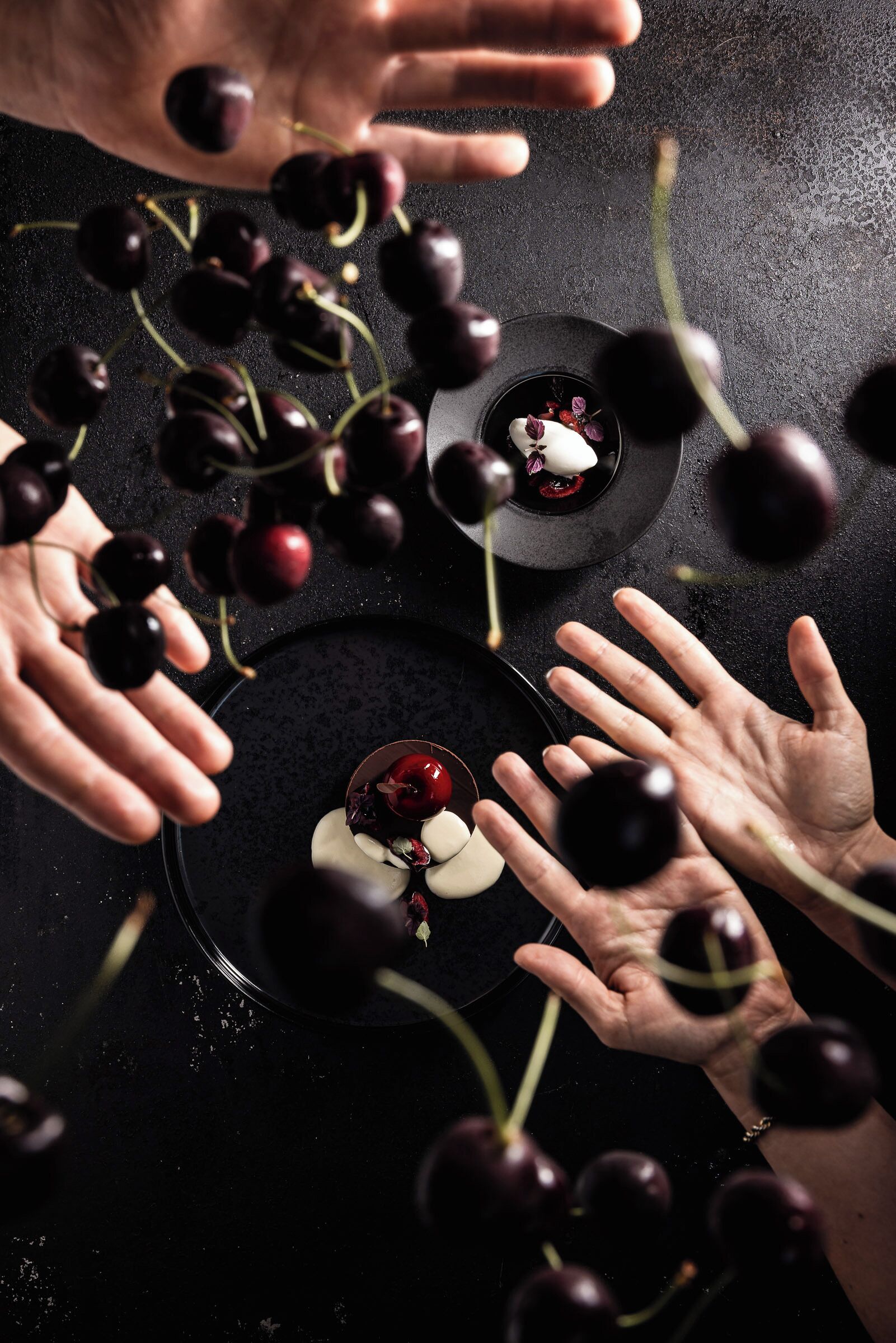 A top-down view of a dining table with a black surface, surrounded by hands reaching towards cherries and a small dish with a cherry on top.