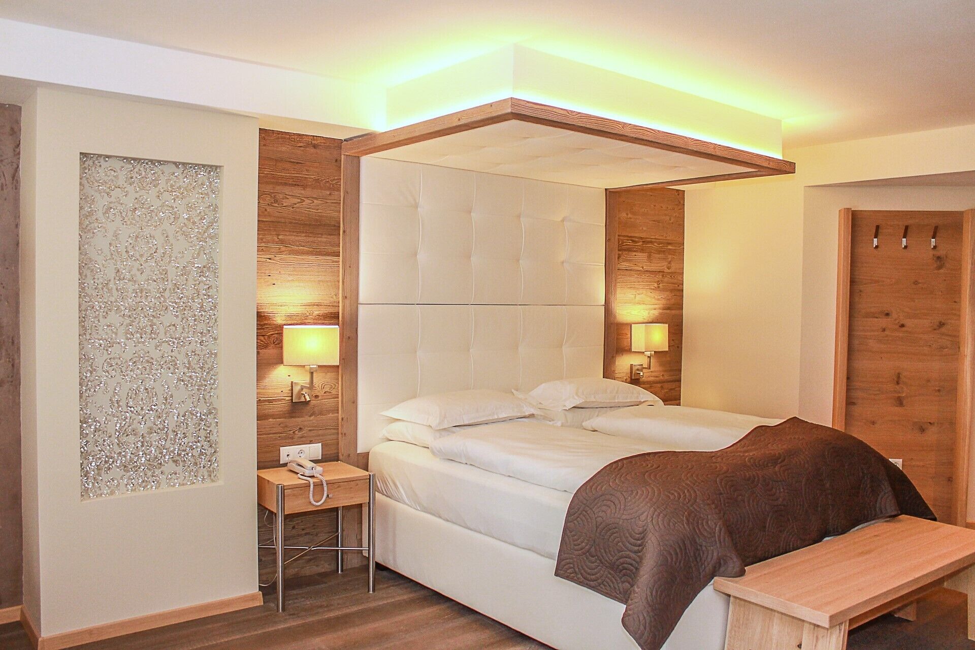 A modern bedroom with a tufted headboard, bedside lamps, a wooden bedside table with a telephone, and a bench at the foot of the bed.