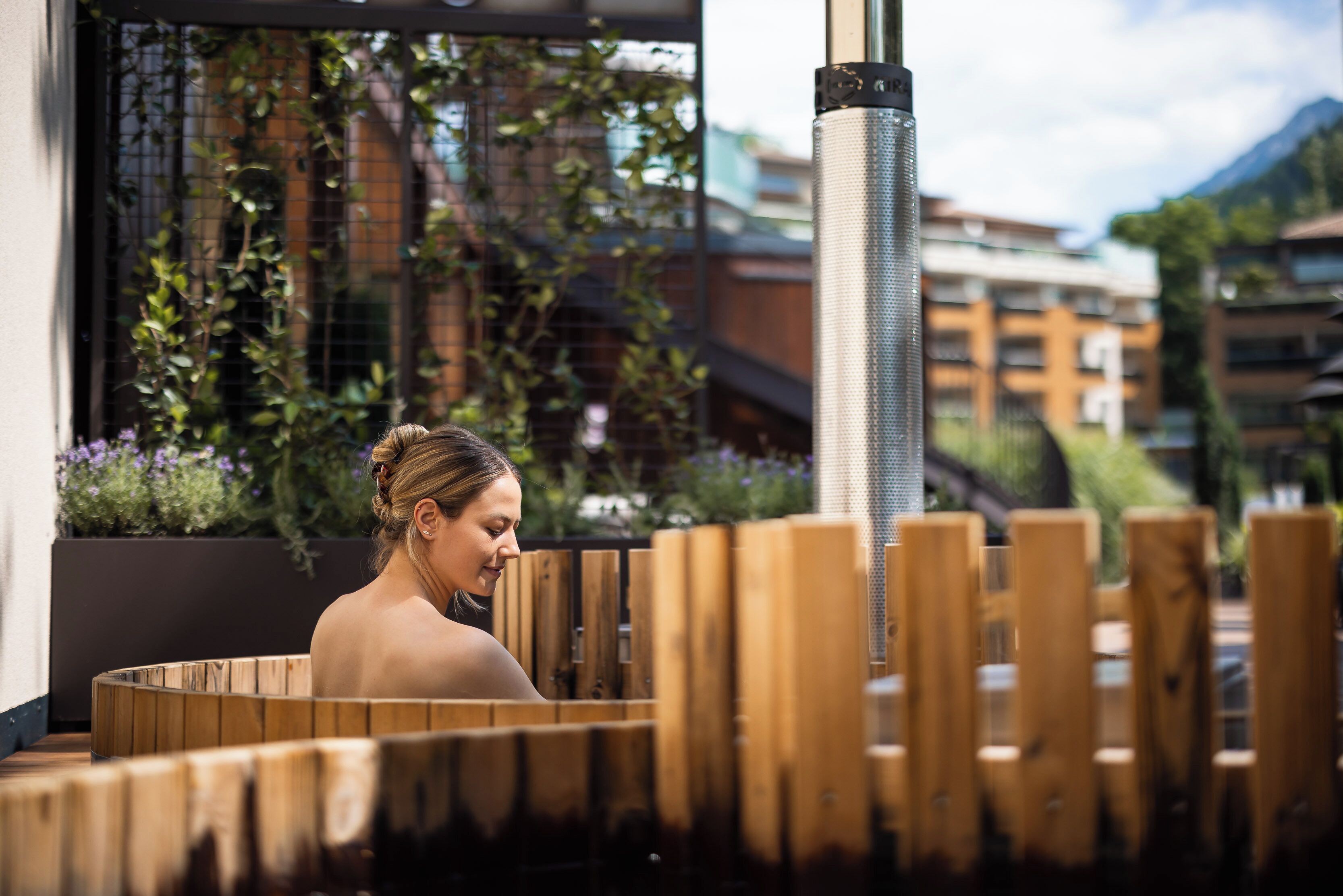 A spa area with wooden benches, a metal column, and a view of buildings and greenery.