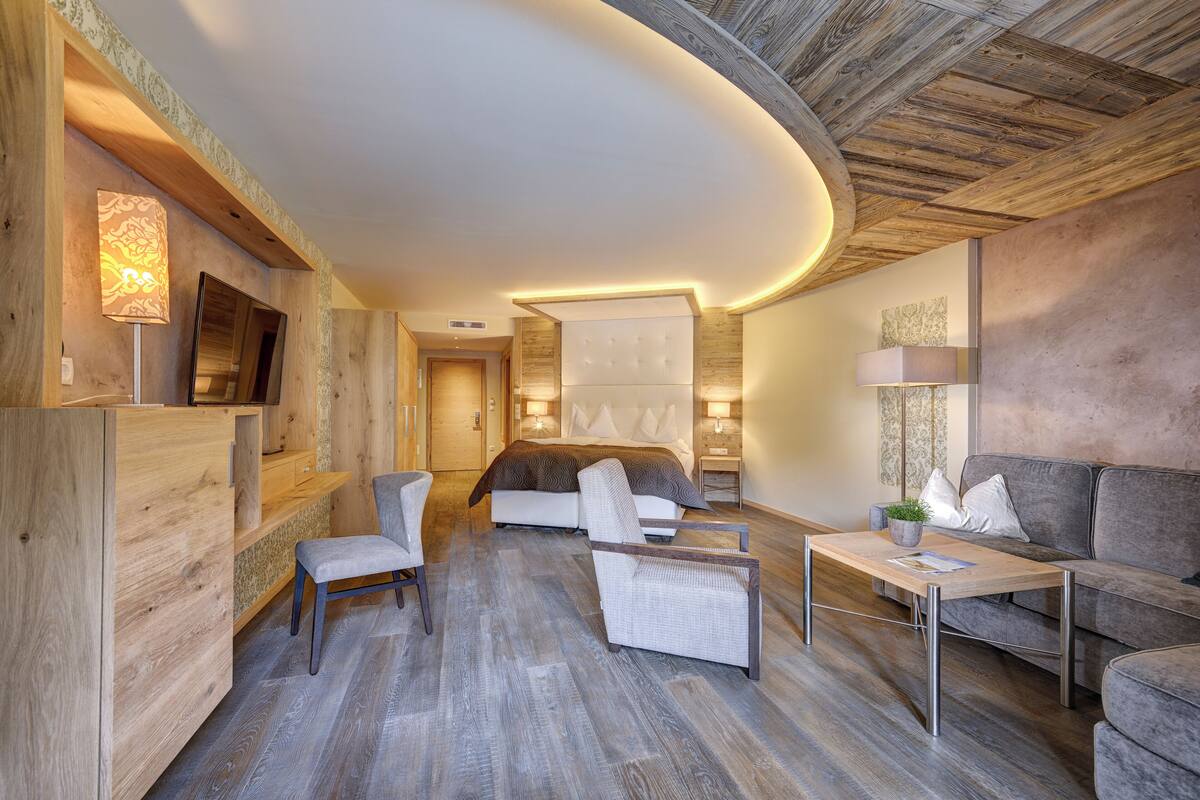 Une chambre d’hôtel moderne avec un plafond en bois, un grand lit avec une tête de lit, deux fauteuils, un canapé, une petite table et un téléviseur fixé au mur.