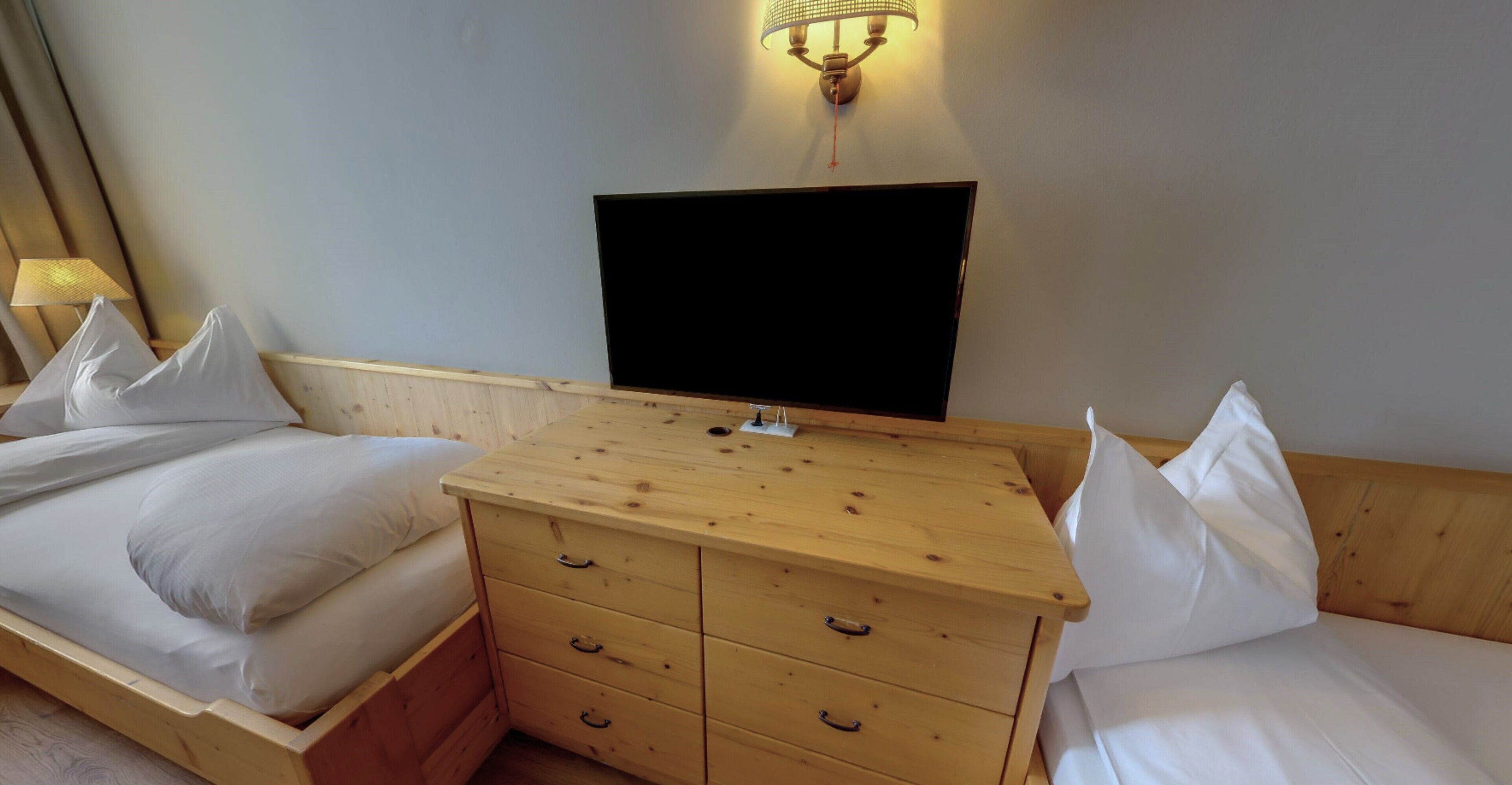 A bedroom with a wooden dresser, two beds, a flat-screen TV, and a wall-mounted lamp.