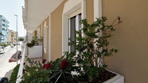 Un balcón de hotel con vista a la calle, una maceta con flores rojas y un cielo despejado.