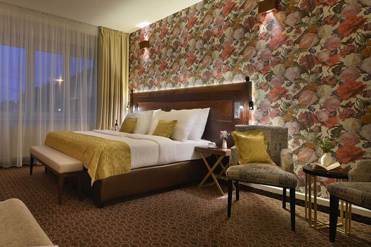 A hotel room with a large bed, a sitting area with a sofa and armchairs, a small table, and a window with curtains.