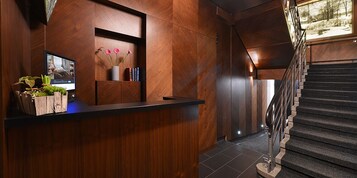 Un área de recepción con un mostrador de madera, un televisor, un jarrón con flores y una escalera que conduce a un piso superior.