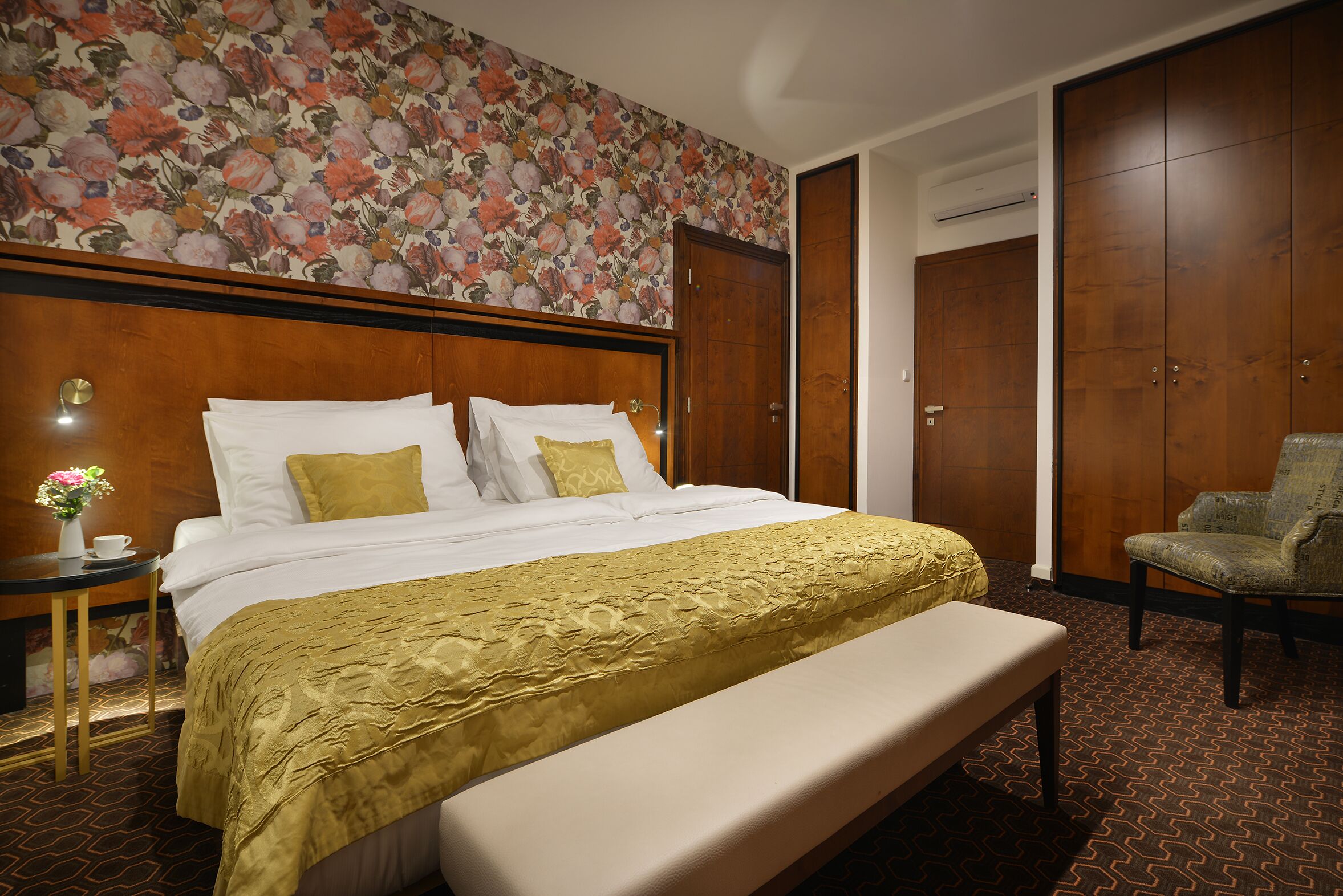 A hotel room with a large bed, a bench, a chair, a nightstand with a vase of flowers, and a wardrobe.