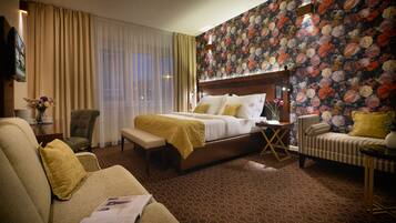 A hotel room with a large bed, a sofa, a chair, and a desk with a lamp.