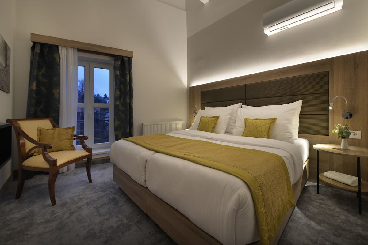 A modern hotel room with a large bed, a wooden chair, a bedside table, and a window with curtains.