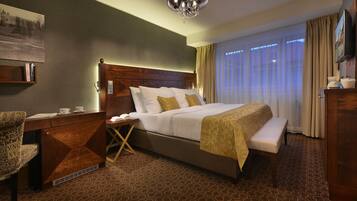 A hotel room with a large bed, a wooden headboard, a bedside table with a lamp, a chair, a bench, and a large window with curtains.
