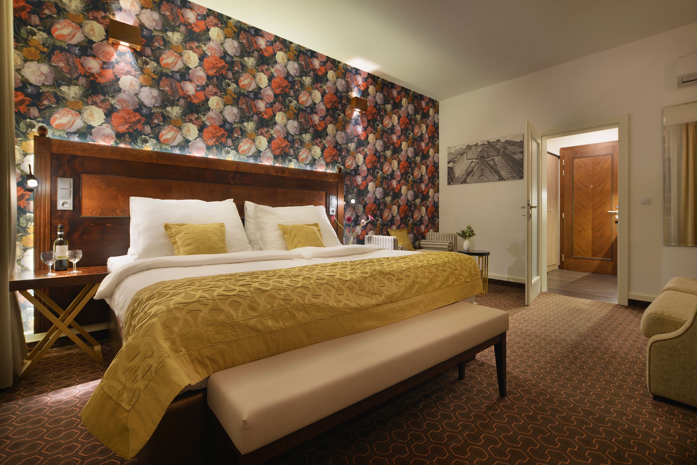 Ein Hotelzimmer mit einem großen Bett, einer Bank, floralem Wandpapier und einer Tür zu einem anderen Raum.