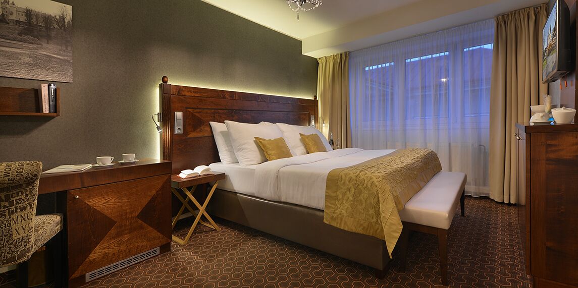A hotel room with a large bed, a wooden headboard, a bedside table with a lamp, a chair, and a desk with a computer.