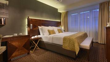 A hotel room with a large bed, a wooden headboard, a bedside table with a lamp, a chair, and a desk with a computer.
