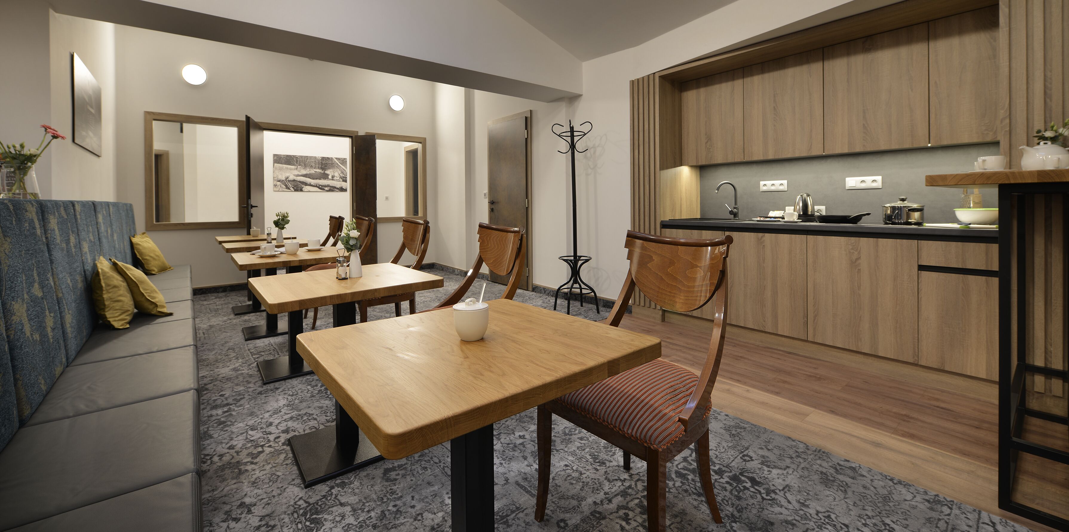 Un espace repas moderne avec des tables et des chaises en bois, une cuisine dotée de meubles en bois et un grand miroir au mur.